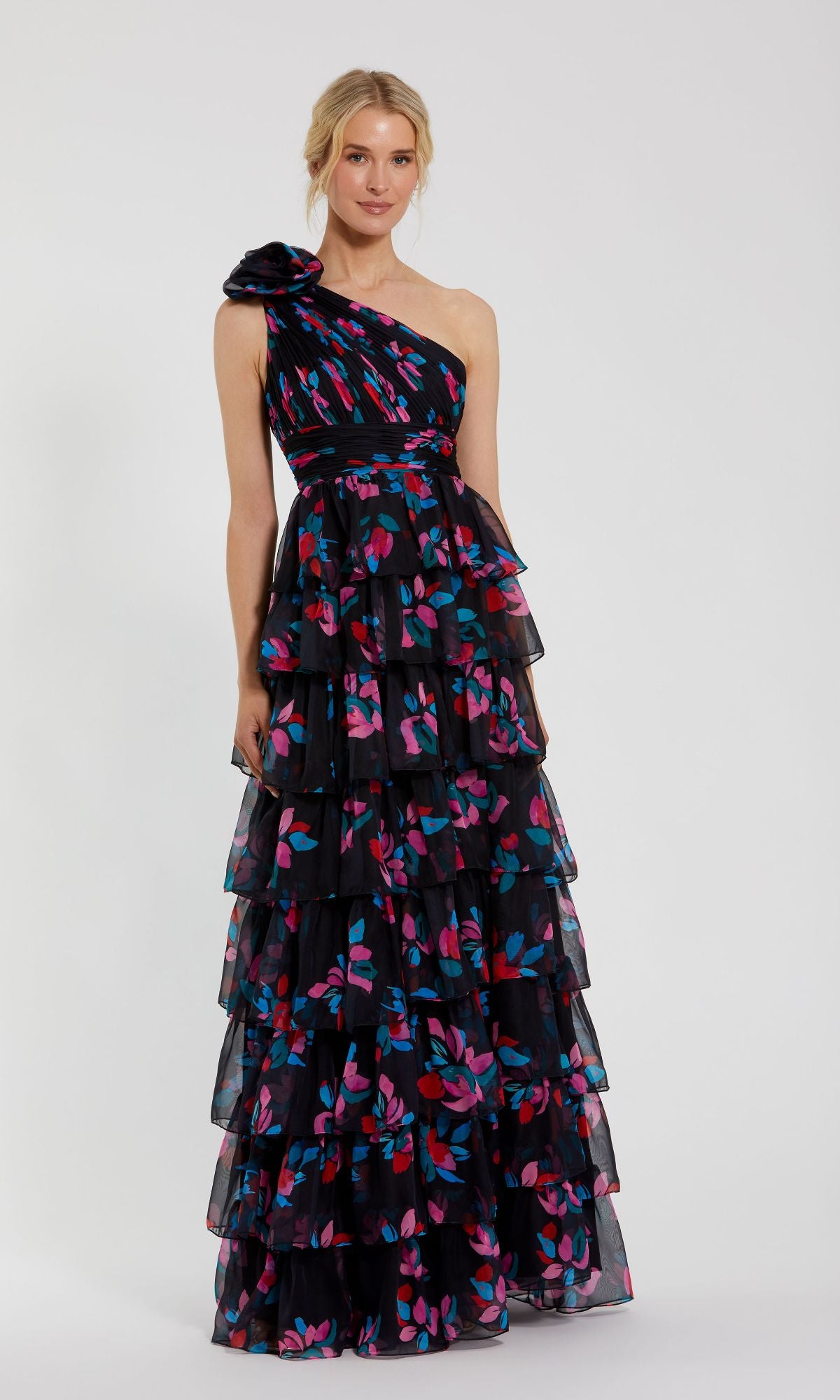 A woman wears the Mac Duggal 56120 Long Print Ruffled Formal Dress—a black, one-shoulder gown with pink and blue floral print and tiered ruffles—standing against a plain background.