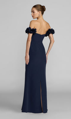 A woman stands with her back to the camera, wearing the Mac Duggal 56131 Floral-Straps Long Formal Dress in navy blue—a graceful off-the-shoulder crepe gown with floral sleeve details, ideal for refined special occasions.