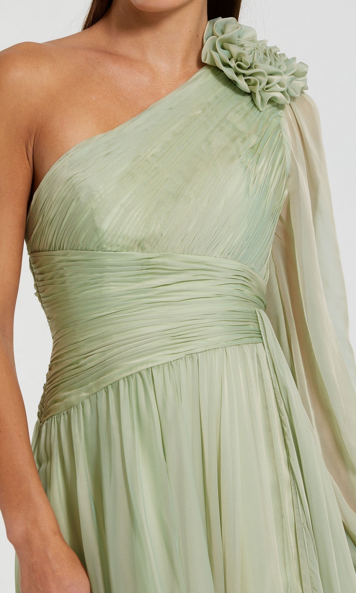 A woman wearing the Mac Duggal 56142 long formal dress, featuring light green fabric with ruching, a floral shoulder detail, and a draped, flowing sleeve.
