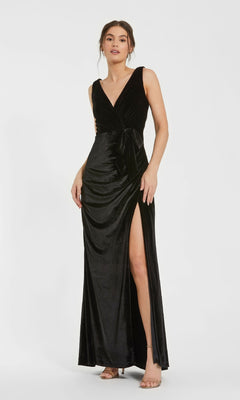A woman stands against a plain white background wearing the Mac Duggal 56144 long black velvet formal dress with a deep V-neck, waist tie, and high side slit.