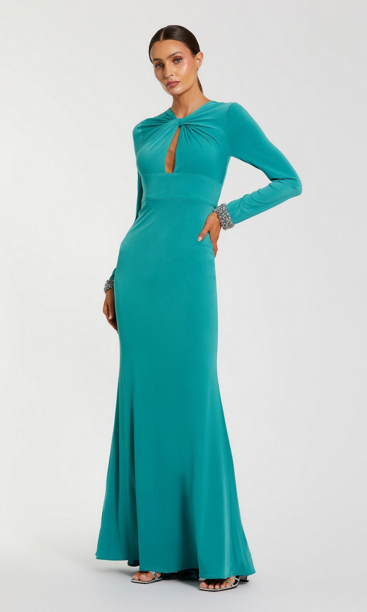 A woman stands against a plain white background wearing the Mac Duggal 56150 long formal dress—a floor-length, long sleeve evening gown in teal—styled with silver bracelets and silver open-toed heels. The dress features a front keyhole cutout.