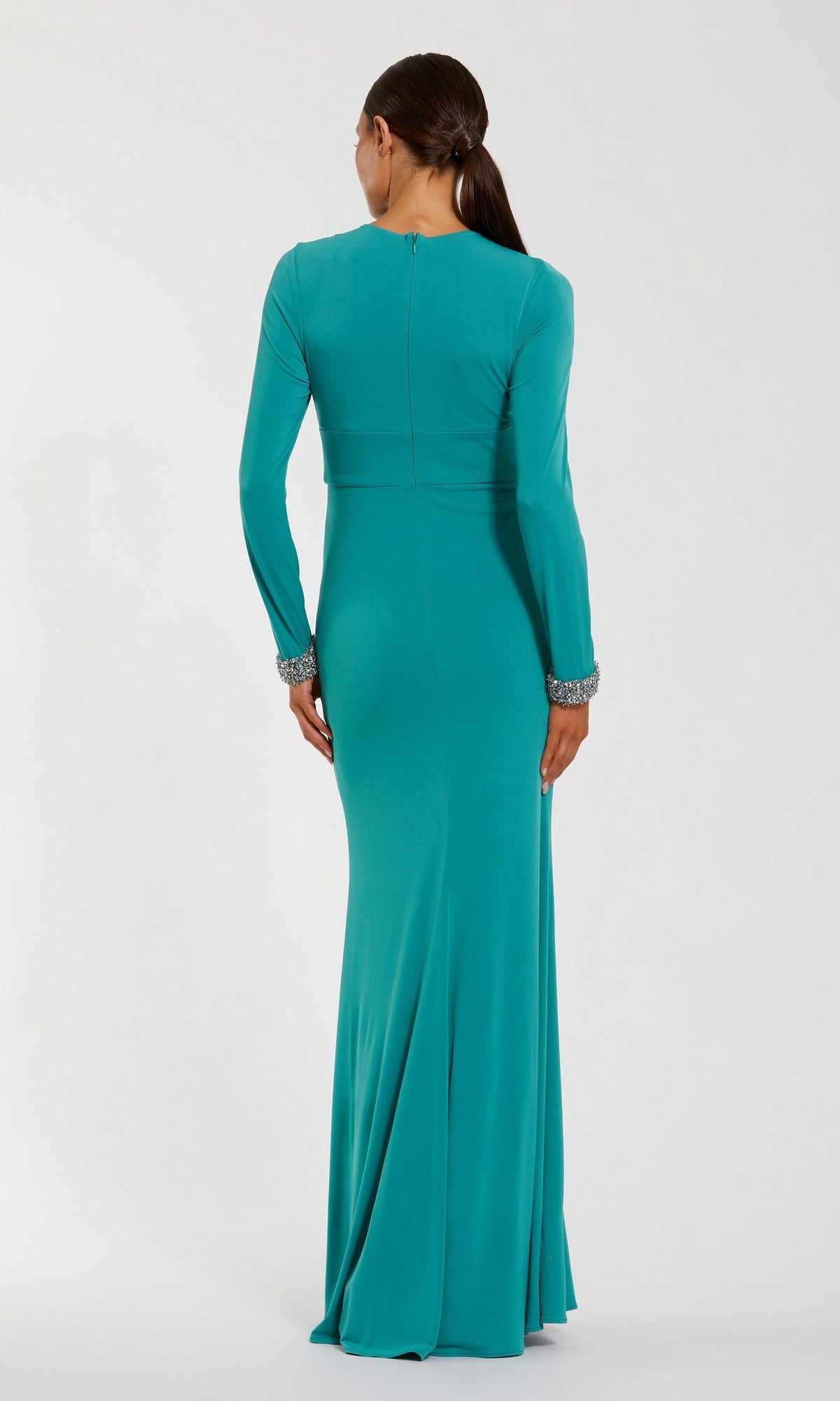 A woman stands with her back to the camera against a plain background, wearing the Mac Duggal 56150 long formal dress—a floor-length teal jersey gown with long sleeves and jeweled cuffs.