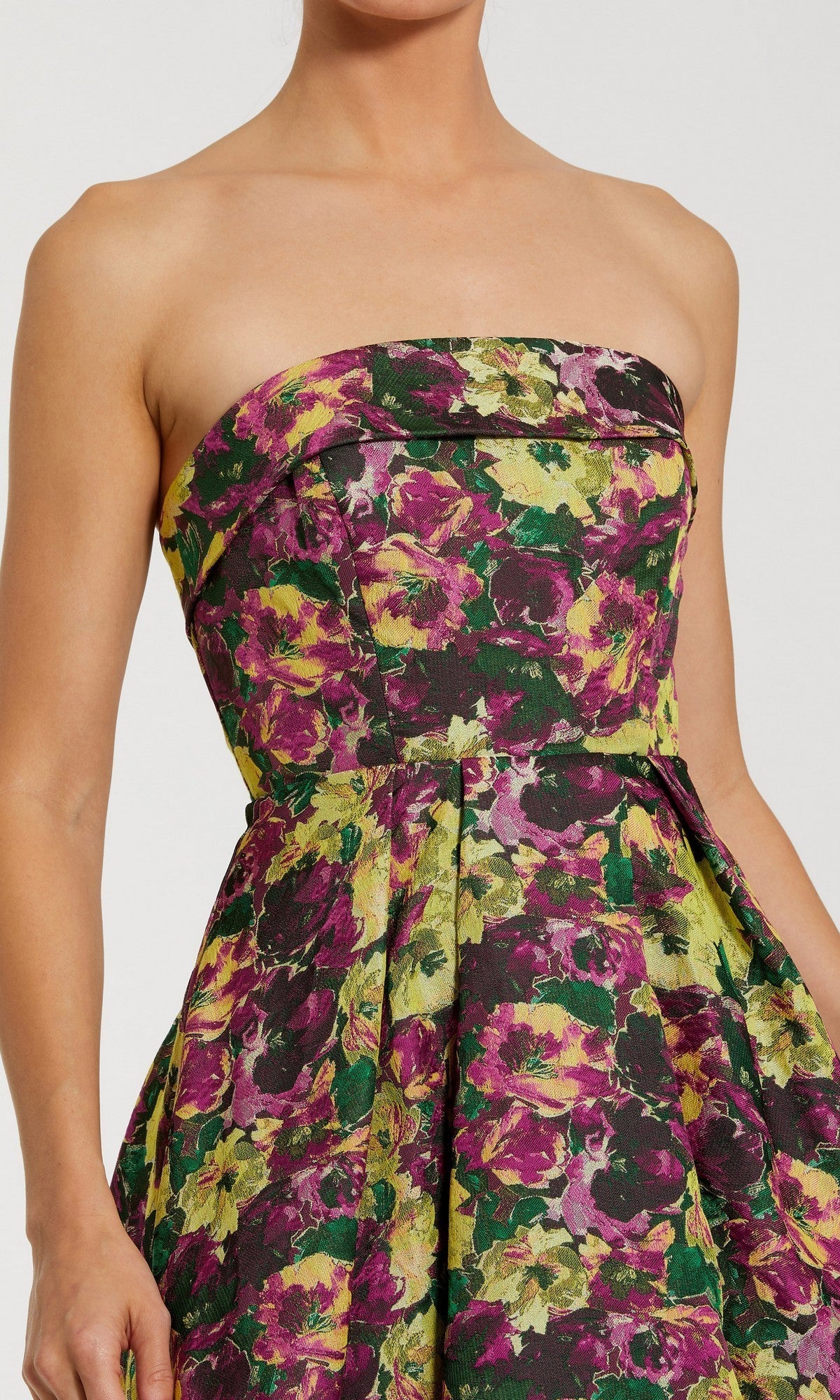 A person wearing the Mac Duggal 56165 Tea-Length Party Dress, featuring a strapless, pleated design with a vibrant floral pattern in shades of purple, green, and yellow. Only the upper body is visible.