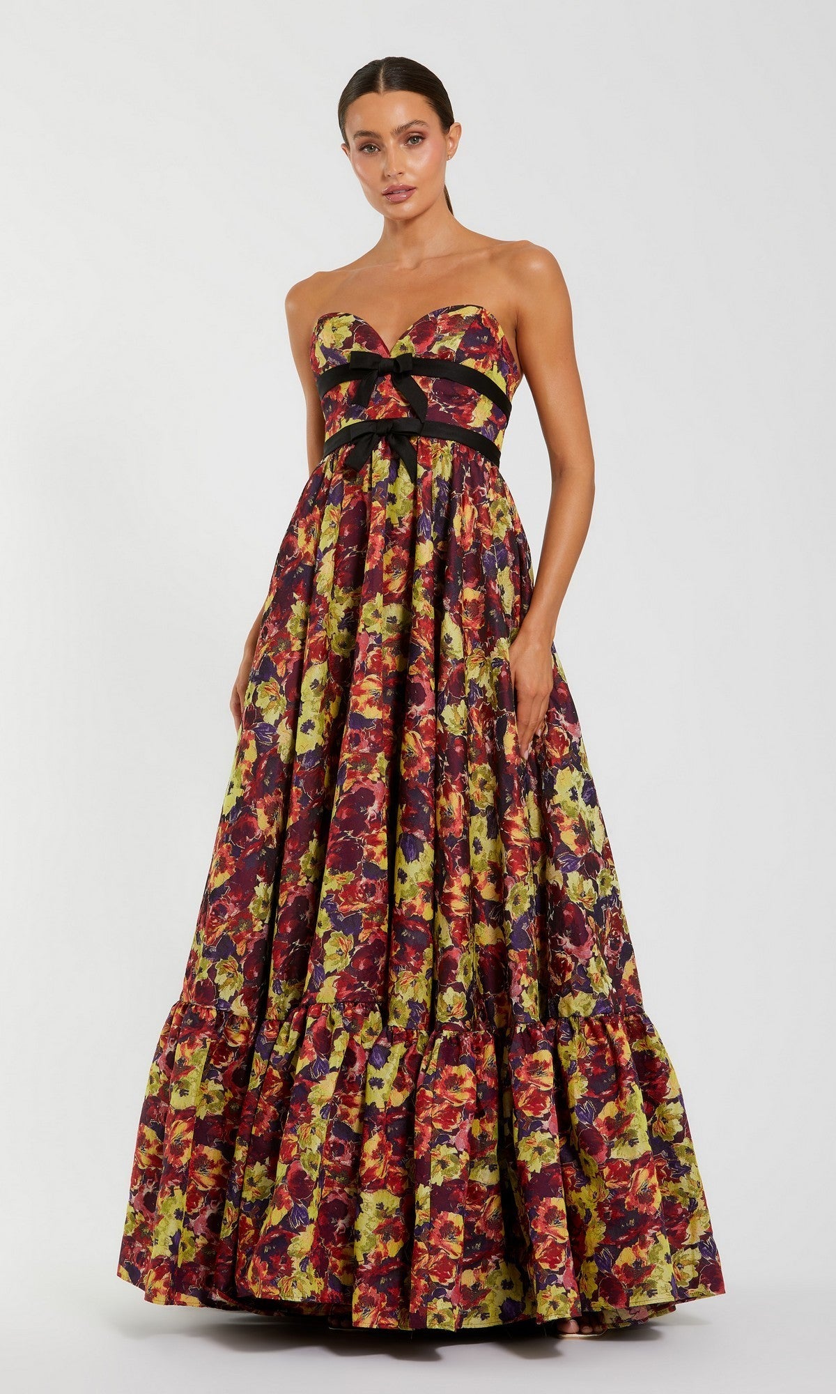 A woman wearing the Mac Duggal 56166 long formal dress in Burgundy Multi—a strapless brocade gown featuring a black bow and a vibrant floral pattern—stands against a plain light background.