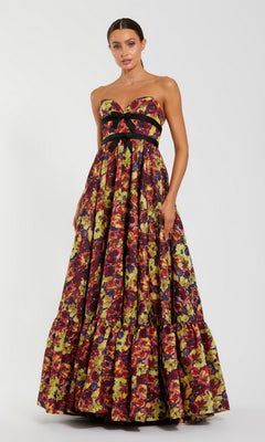A woman wearing the Mac Duggal 56166 long formal dress in Burgundy Multi—a strapless brocade gown featuring a black bow and a vibrant floral pattern—stands against a plain light background.