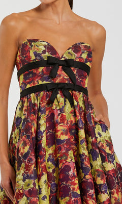 A woman wears the “Long Formal Dress: Mac Duggal 56166,” a strapless brocade formal gown showcasing a floral pattern, adorned with a sweetheart neckline and elegant black ribbon bows across the bodice.