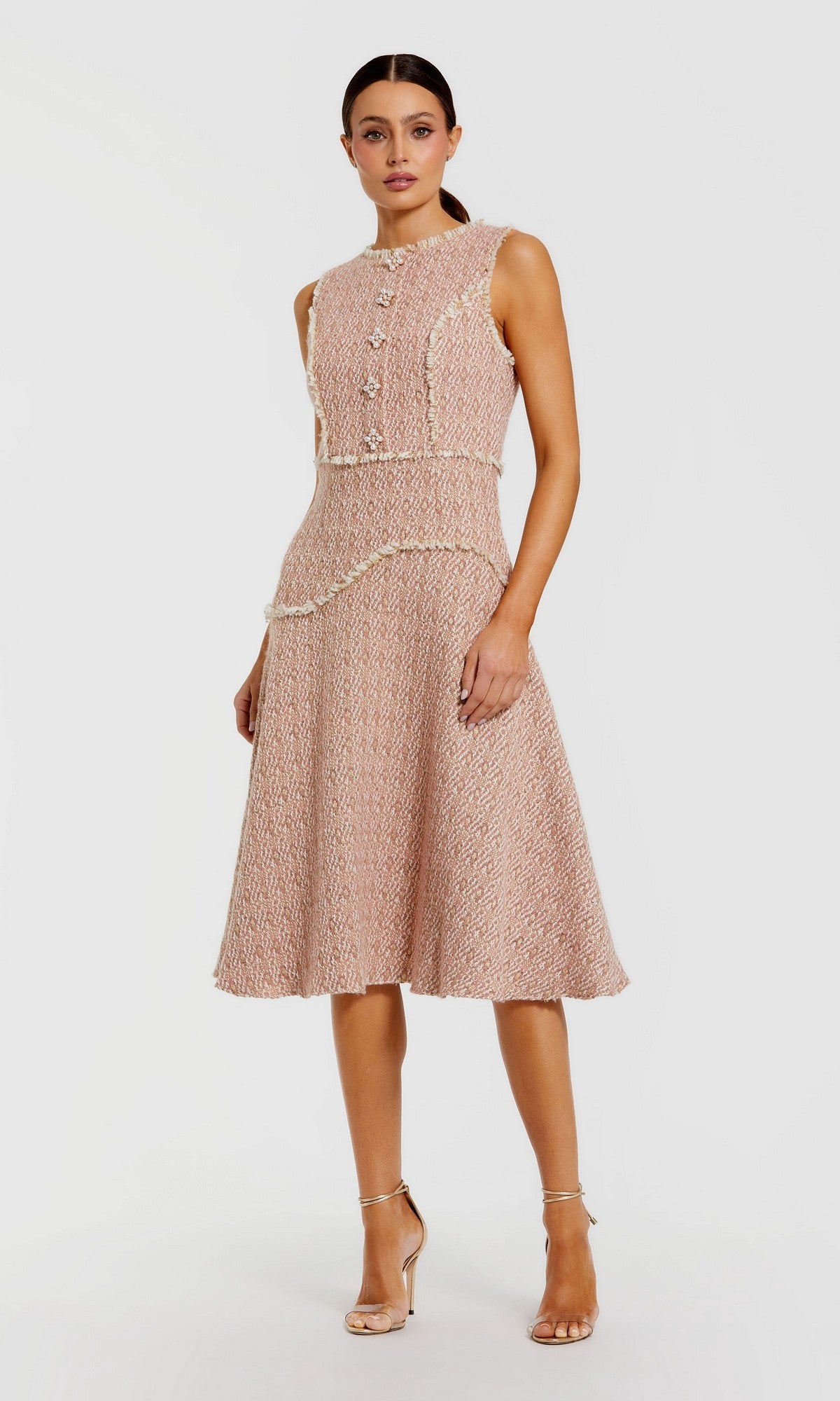 A woman stands against a plain white background, wearing the Mac Duggal 56172 Button-Up Boucle Midi Dress—a sleeveless, knee-length textured pink dress featuring a fitted waist, decorative faux pearl buttons, cream trim—and nude high-heeled sandals.