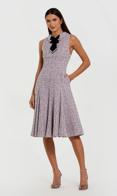 A woman models the Mac Duggal 56175 short wedding guest dress—sleeveless, knee-length, lilac boucle with a black collar bow—paired with silver high-heeled sandals against a plain backdrop.