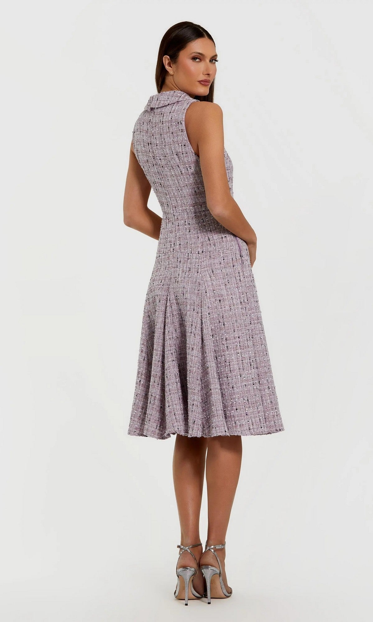 A woman stands looking over her shoulder, wearing the Mac Duggal 56175 short wedding guest dress—sleeveless, knee-length, lilac boucle with a flared skirt—paired with silver high-heeled sandals.