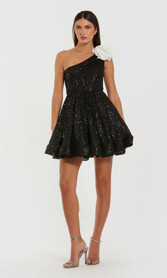 A woman wears the Mac Duggal 56179 short semi-formal dress in black ivory with a one-shoulder design and large white floral detail, paired with silver high-heeled sandals.
