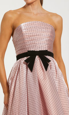 A woman wearing the Mac Duggal 56184 long formal dress: a strapless organza gown in light pink with black polka dots, featuring a fitted bodice and accented with a black velvet bow at the waist.