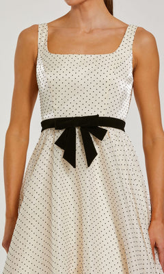 A woman wearing the Mac Duggal 56185 long formal dress—a sleeveless white organza A-line gown adorned with black polka dots and finished with a black bow-tied belt at the waist—channels a playful yet sophisticated polka-dot style.