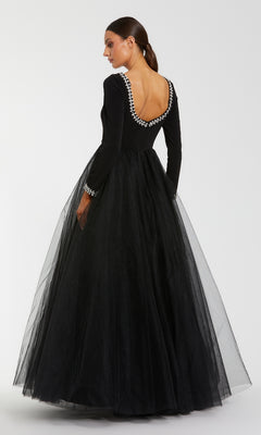 A woman poses in the Mac Duggal 56186, a long sleeve black formal gown featuring a tulle skirt, deep scoop back, and pearl accents on the neckline and cuffs.