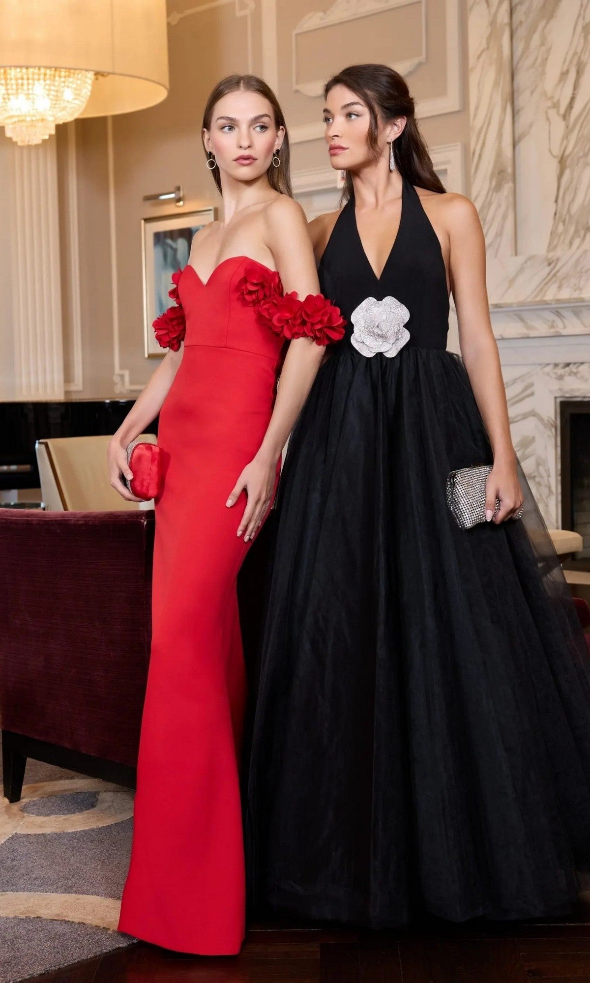 Two women in formal gowns stand indoors; one wears the Long Formal Dress: Mac Duggal 56187, a fitted red dress with floral accents, while the other stuns in a black halter ballgown with a white flower detail at the waist.