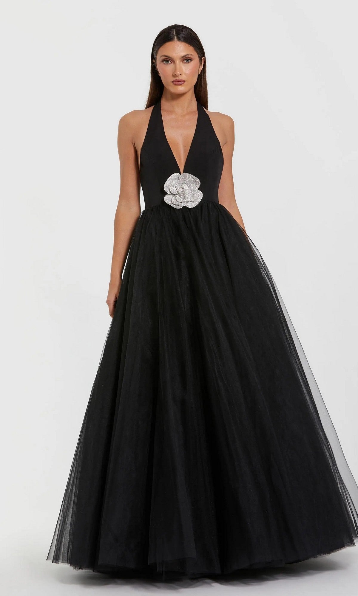 A woman stands against a plain white background wearing the Long Formal Dress: Mac Duggal 56187, a floor-length gown featuring a deep plunging neckline and a large white flower embellishment at the waist.