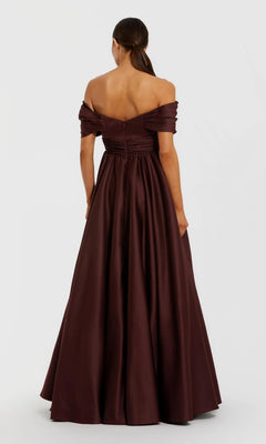 A woman with her hair in a low ponytail faces away, wearing the Mac Duggal 56188 Long Formal Dress—an off-the-shoulder, floor-length dark burgundy satin gown with a fitted bodice and flared skirt. Plain white background.