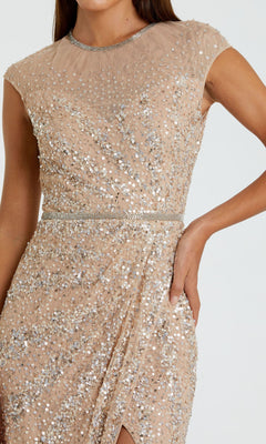 A person wearing the Mac Duggal 5619 Sleeveless Beaded Long Formal Dress, featuring a fitted waist, draped skirt, and elegant nude-toned sequins, stands with one hand on their hip.