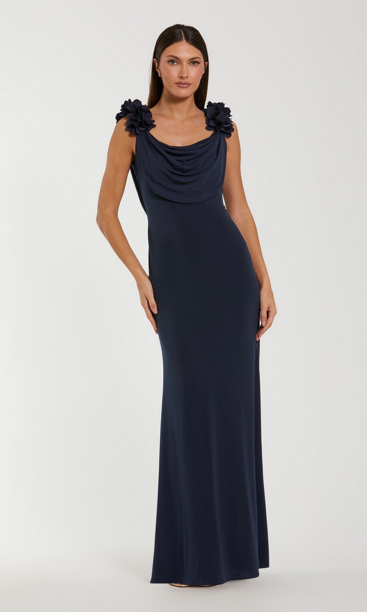 A woman wearing the Mac Duggal 56195 Long Formal Dress in Midnight—a floor-length, sleeveless jersey gown featuring a draped neckline and floral shoulder accents—stands against a plain light background.