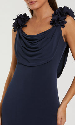 A woman wearing the Mac Duggal 56195 long formal dress—a navy blue sleeveless gown featuring a draped neckline and large floral appliqués on the shoulders, styled in an elegant silhouette.