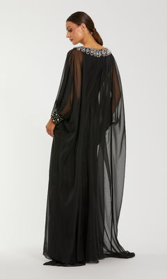 A woman wears the Mac Duggal 56203 long formal dress, featuring black chiffon fabric and rhinestone-embellished neckline and cuffs, as she stands against a white background, facing slightly away from the camera.