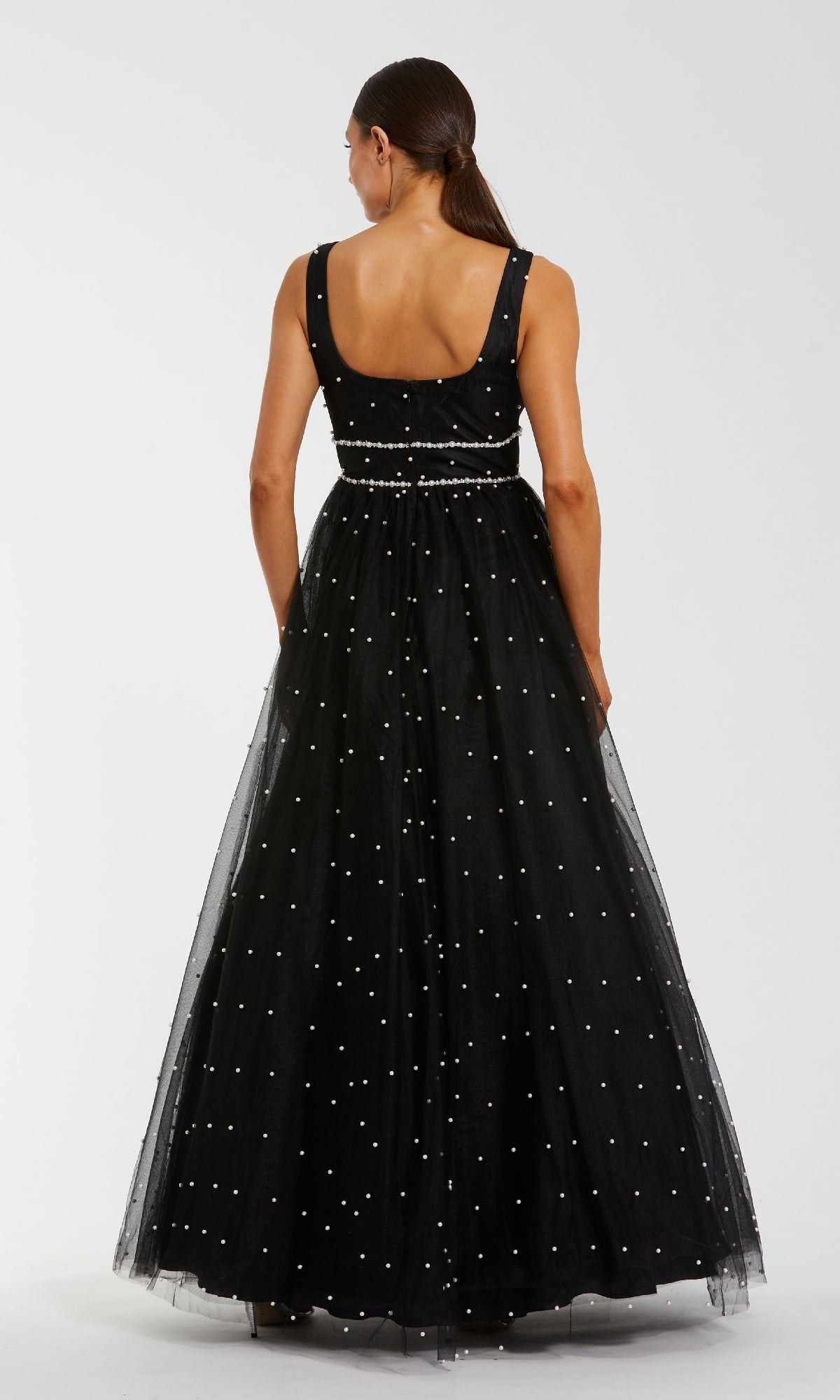A woman with long brown hair in a low ponytail wears the Mac Duggal 56204 long formal dress—an A-line black ball gown with a floor-length tulle skirt and white polka dots—seen from the back against a plain white background.