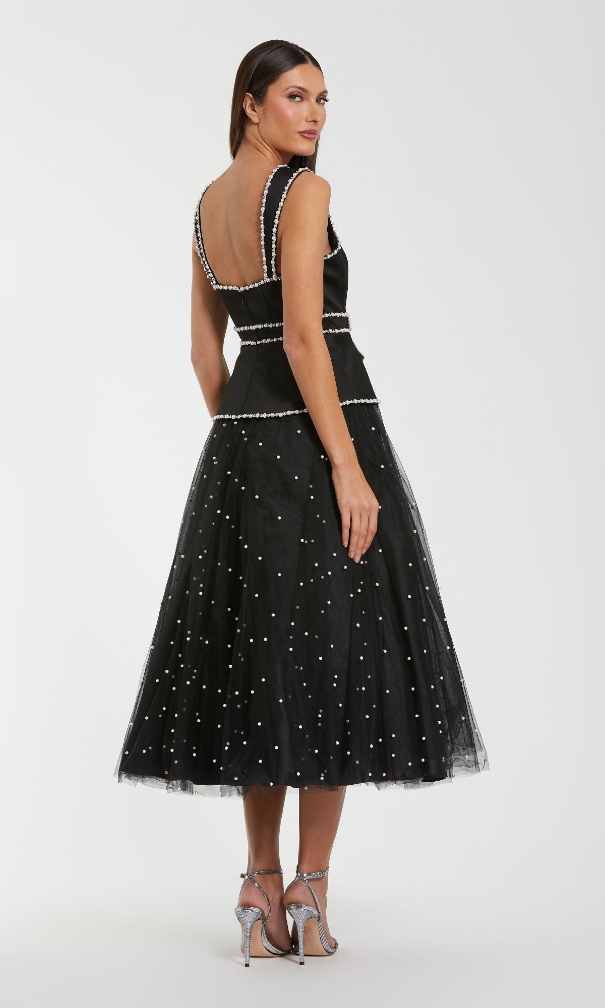 A woman wearing the Mac Duggal 56206 Black Midi Wedding Guest Dress—sleeveless, mid-calf tulle with white pearl accents—looks over her shoulder, styled elegantly with silver heels against a plain backdrop.