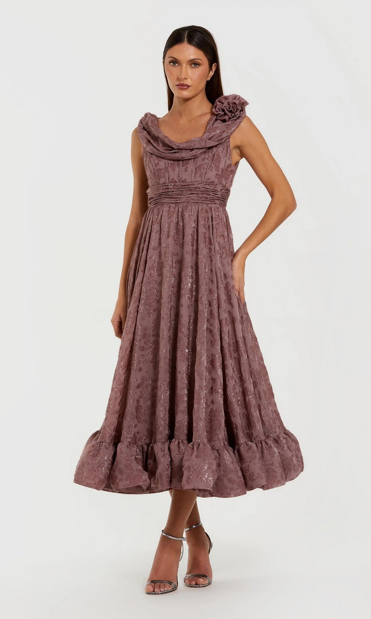 A woman poses in a mauve Mac Duggal 56223 midi semi-formal dress with textured fabric, ruffled hem, and floral one-shoulder detail, paired with metallic heels against a plain background.
