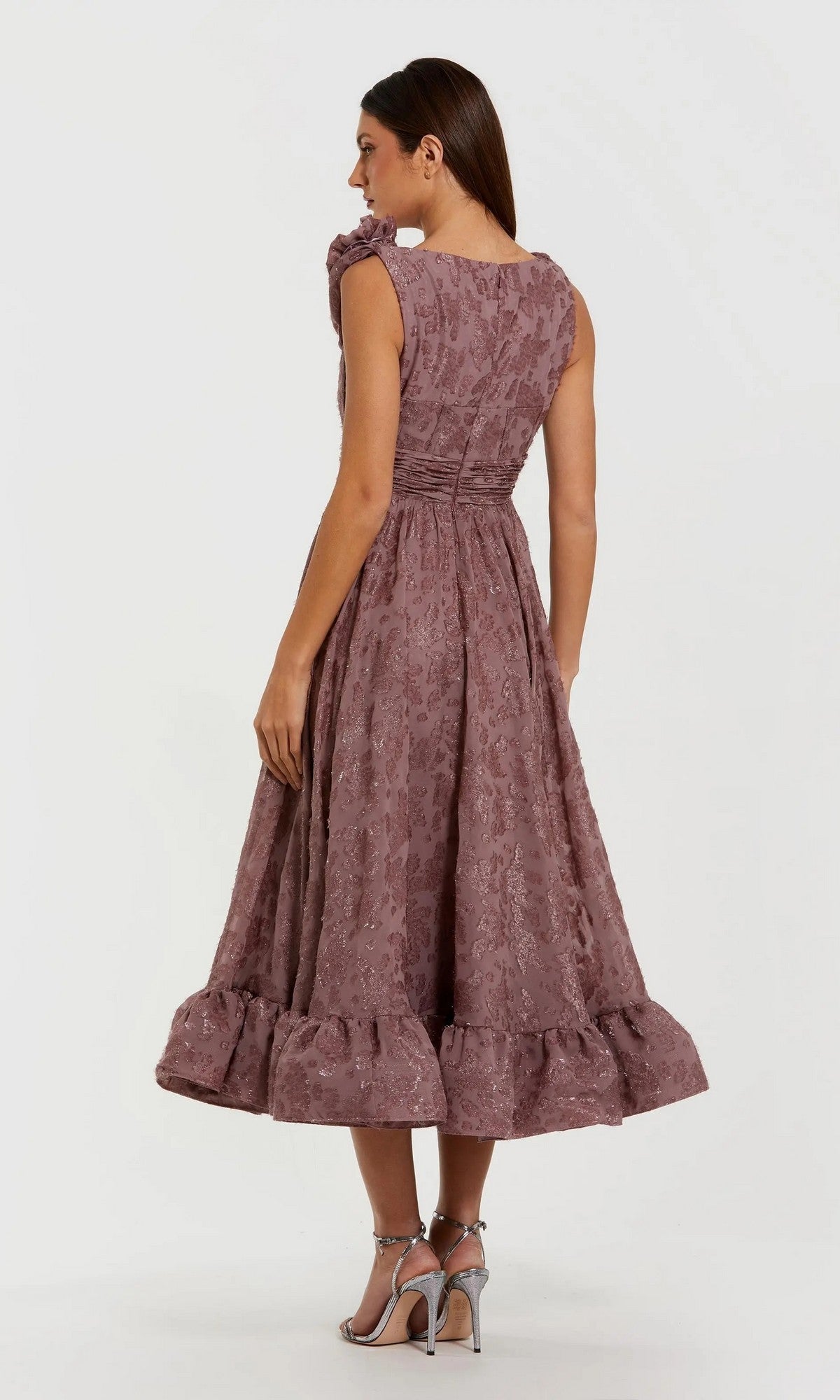A woman stands facing away, wearing the Midi Semi-Formal Dress: Mac Duggal 56223 in mauve with a ruffled hem and floral texture, paired with silver high-heeled sandals.