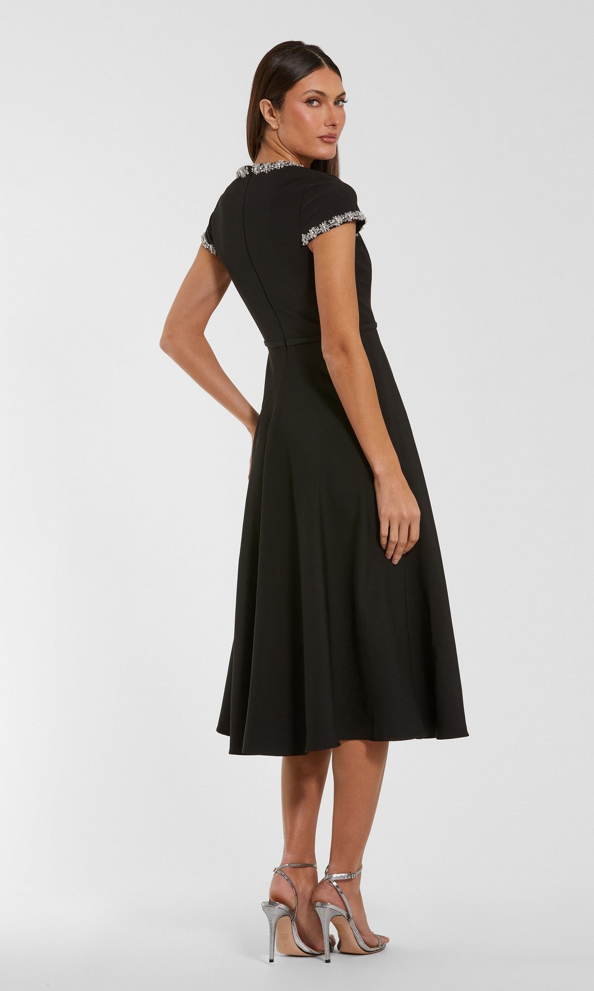 A woman wearing the Mac Duggal 56242 midi party dress in black, featuring short sleeves and embellished trim, paired with silver high-heeled sandals, stands against a plain background.