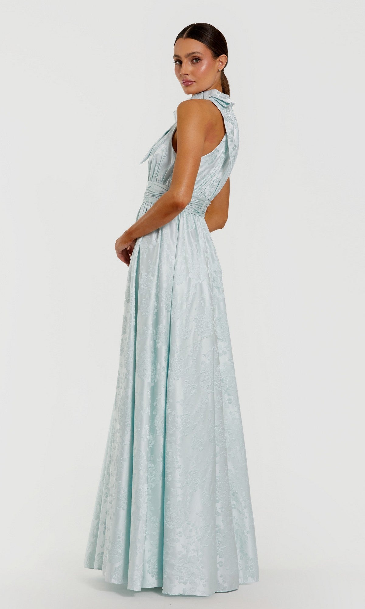 A woman poses slightly to the side against a plain white background, wearing a light blue, floor-length sleeveless evening gown with a high neckline and textured brocade fabric. The formal dress is the Long Formal Dress: Mac Duggal 56246, radiating sophistication and timeless elegance.