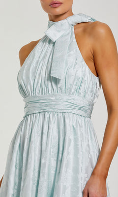 A woman wearing the Long Formal Dress: Mac Duggal 56246, featuring a pale blue floral jacquard fabric, a wide tied halter neckline, and a ruched waistband for an elegant formal evening look.