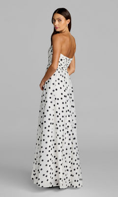 Wearing the Mac Duggal 56248 long formal dress, a woman stands against a plain gray background. The strapless, floor-length gown features bold black polka dots, as she looks over her shoulder.