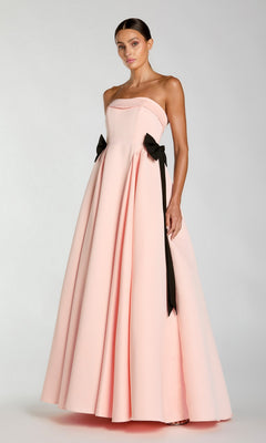 A woman is wearing the Long Formal Dress: Mac Duggal 56251, a floor-length blush satin A-line gown featuring a strapless neckline and two black bows at the waist, standing against a plain light gray background.