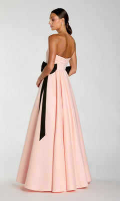 A woman stands against a plain background, facing slightly away, wearing the Mac Duggal 56251: a strapless, floor-length pale pink formal gown with a black ribbon belt.