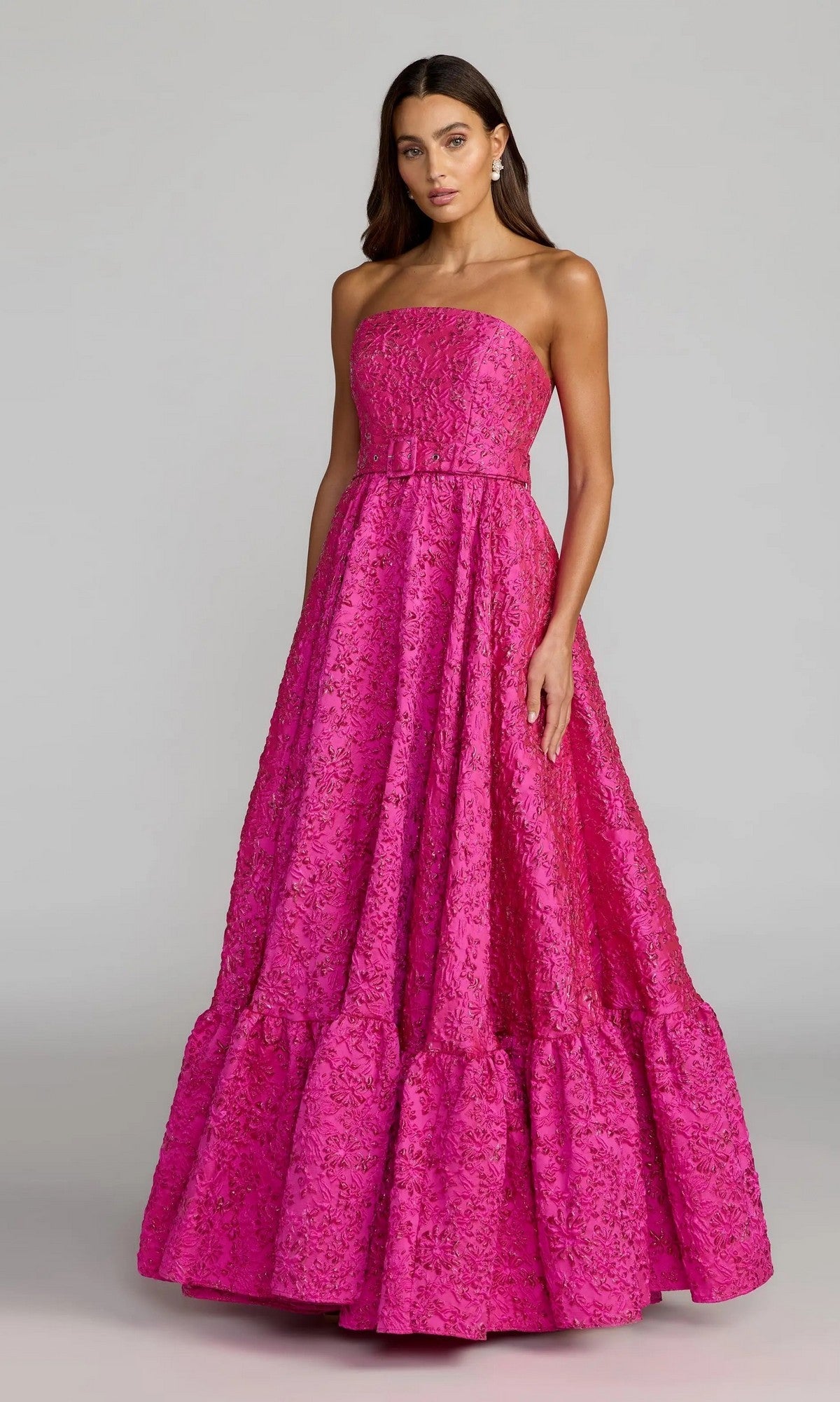 A woman stands against a plain gray background, wearing the Mac Duggal 56277 long formal dress—a strapless, floor-length gown in textured bright pink, featuring a fitted bodice and a ruffled hem.