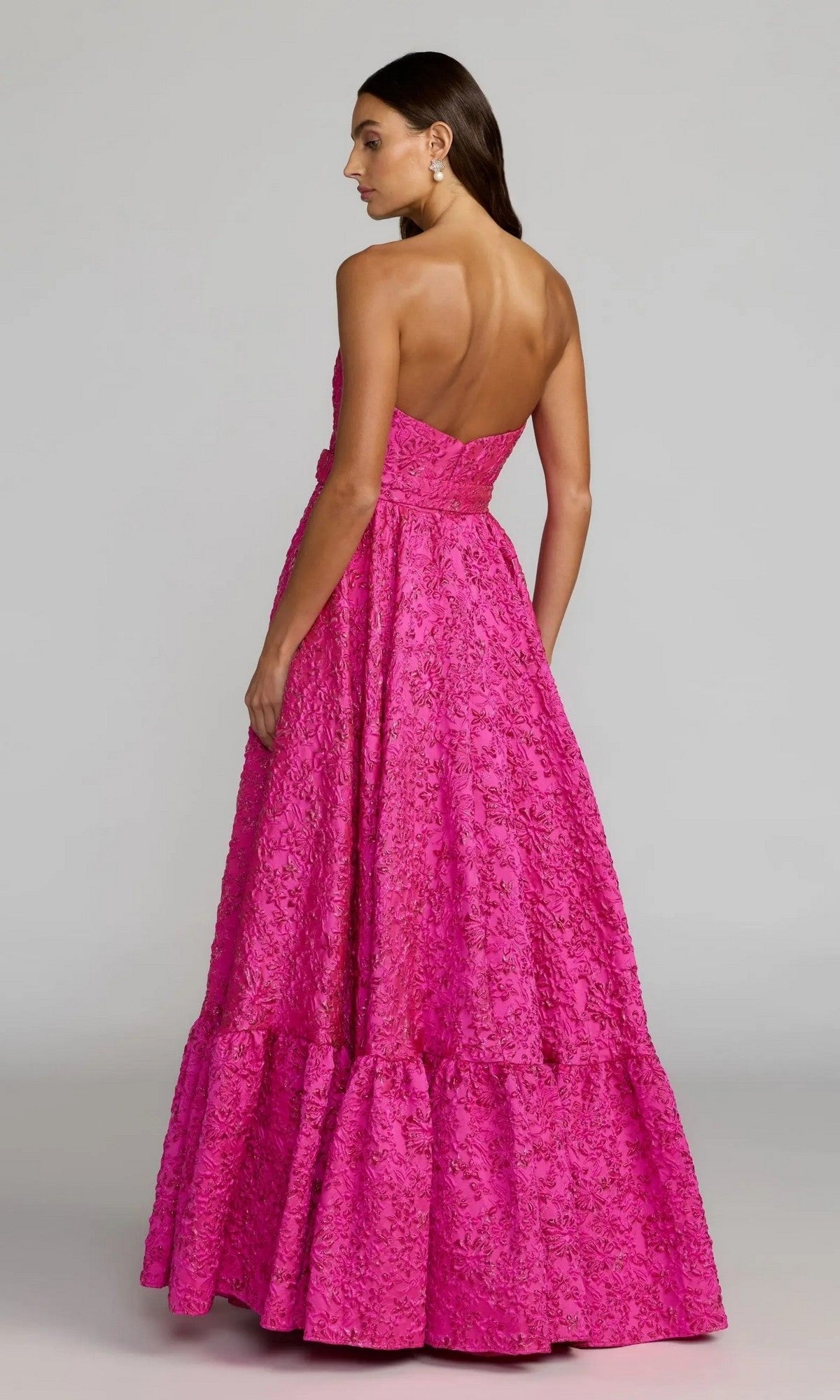 A woman wearing the Mac Duggal 56277 long formal dress—a pink, strapless gown with a floor-length, textured fuchsia tiered skirt reminiscent of a floral brocade ballgown—stands against a plain light background.