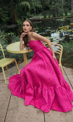 A woman wearing the Mac Duggal 56277 long formal dress, featuring a bright pink textured strapless design, sits at a yellow table and chair set near a pond in a lush garden setting.