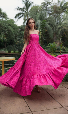 A woman stands outdoors on a patio surrounded by lush greenery, exuding elegance in the Mac Duggal 56277 long formal dress in hot pink, featuring a strapless design with a textured finish and ruffled hem.