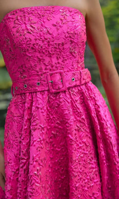 A person wears the Long Formal Dress: Mac Duggal 56277—a strapless, textured pink gown with a fitted waist and matching belt. Reminiscent of a floral brocade ballgown, it features delicate floral embroidery for an elegant finish.