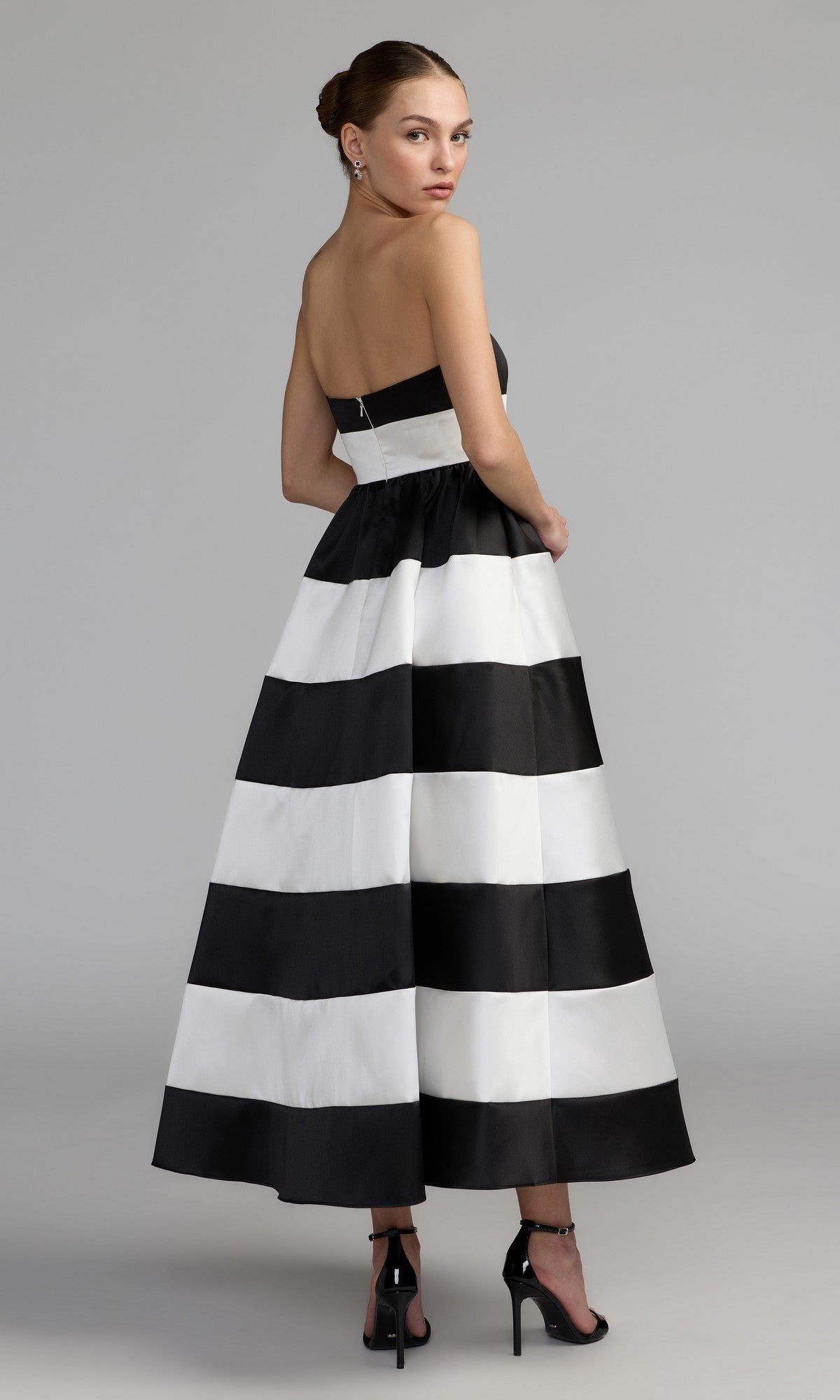 A woman models the Mac Duggal 56281 Tea-Length Party Dress—a strapless design with bold black and white stripes—as she stands with her back slightly turned, wearing black high heels and drop earrings against a plain gray background.
