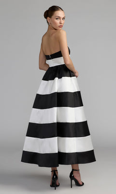 A woman models the Mac Duggal 56281 Tea-Length Party Dress—a strapless design with bold black and white stripes—as she stands with her back slightly turned, wearing black high heels and drop earrings against a plain gray background.