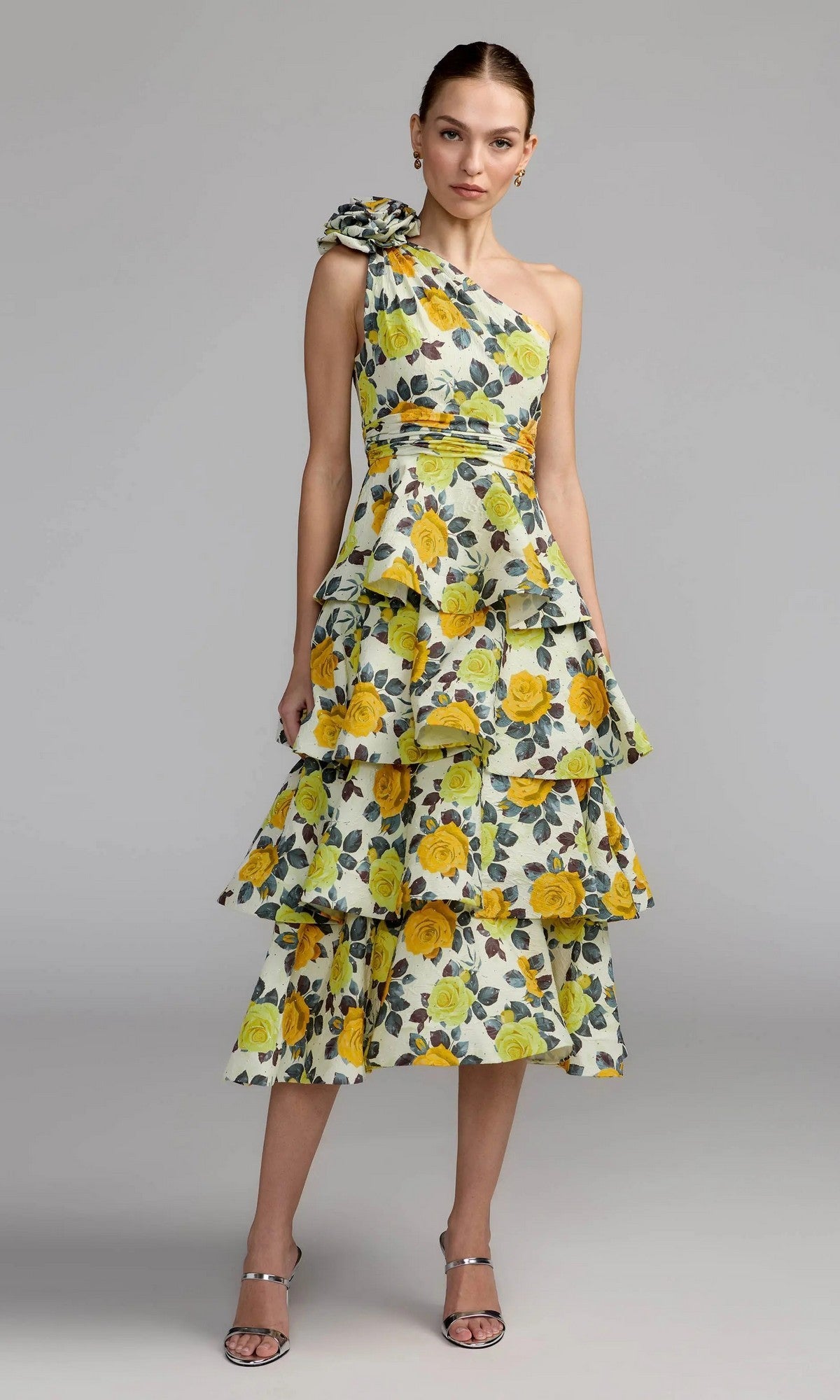 A woman poses against a plain gray background in the Mac Duggal 56314 Midi Party Dress, showcasing its yellow rose floral-printed charmeuse fabric, tiered ruffles, and asymmetrical one-shoulder design. She completes her look with hoop earrings and strappy sandals.