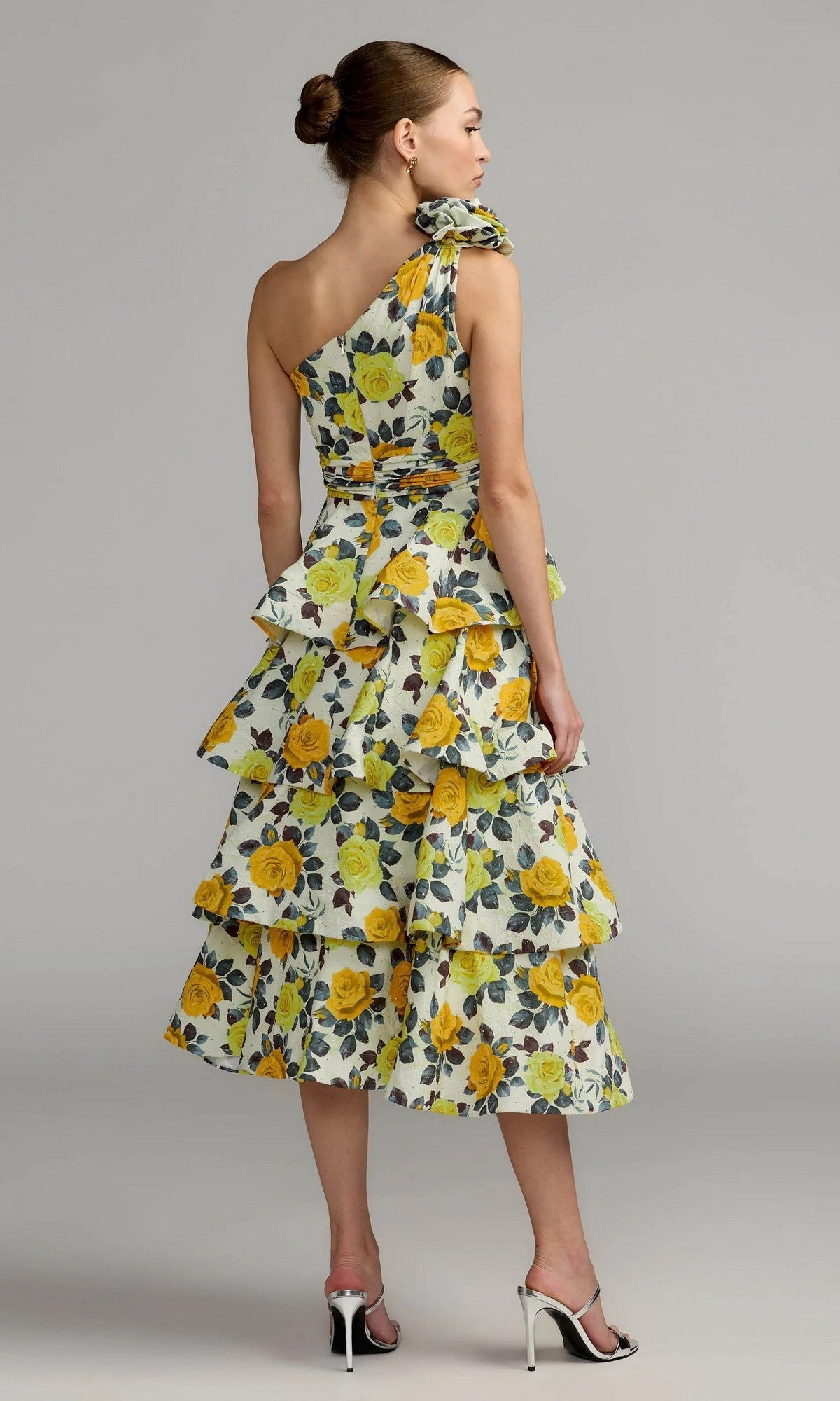 A woman in the Midi Party Dress: Mac Duggal 56314—a yellow one-shoulder midi dress featuring rose print, tiered ruffles, and white heeled sandals—stands against a plain gray background, showcasing a chic look perfect for any occasion.