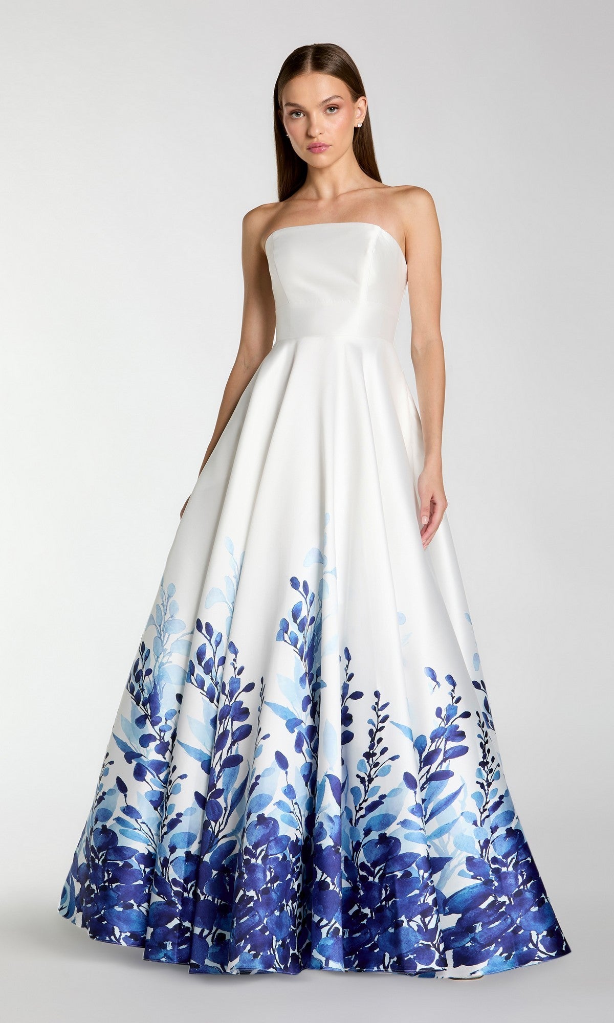 A woman stands gracefully against a plain light background, wearing the Long Formal Dress: Mac Duggal 56316—a stunning strapless ballgown in White Multi, featuring a white silhouette beautifully adorned with a blue floral pattern around the lower half.