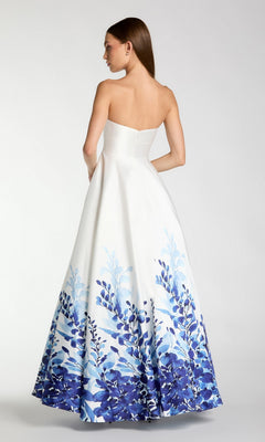 A woman stands with her back to the camera against a plain background, wearing the Mac Duggal 56316 long formal dress—a strapless Mikado ballgown featuring a blue floral pattern along the lower half.