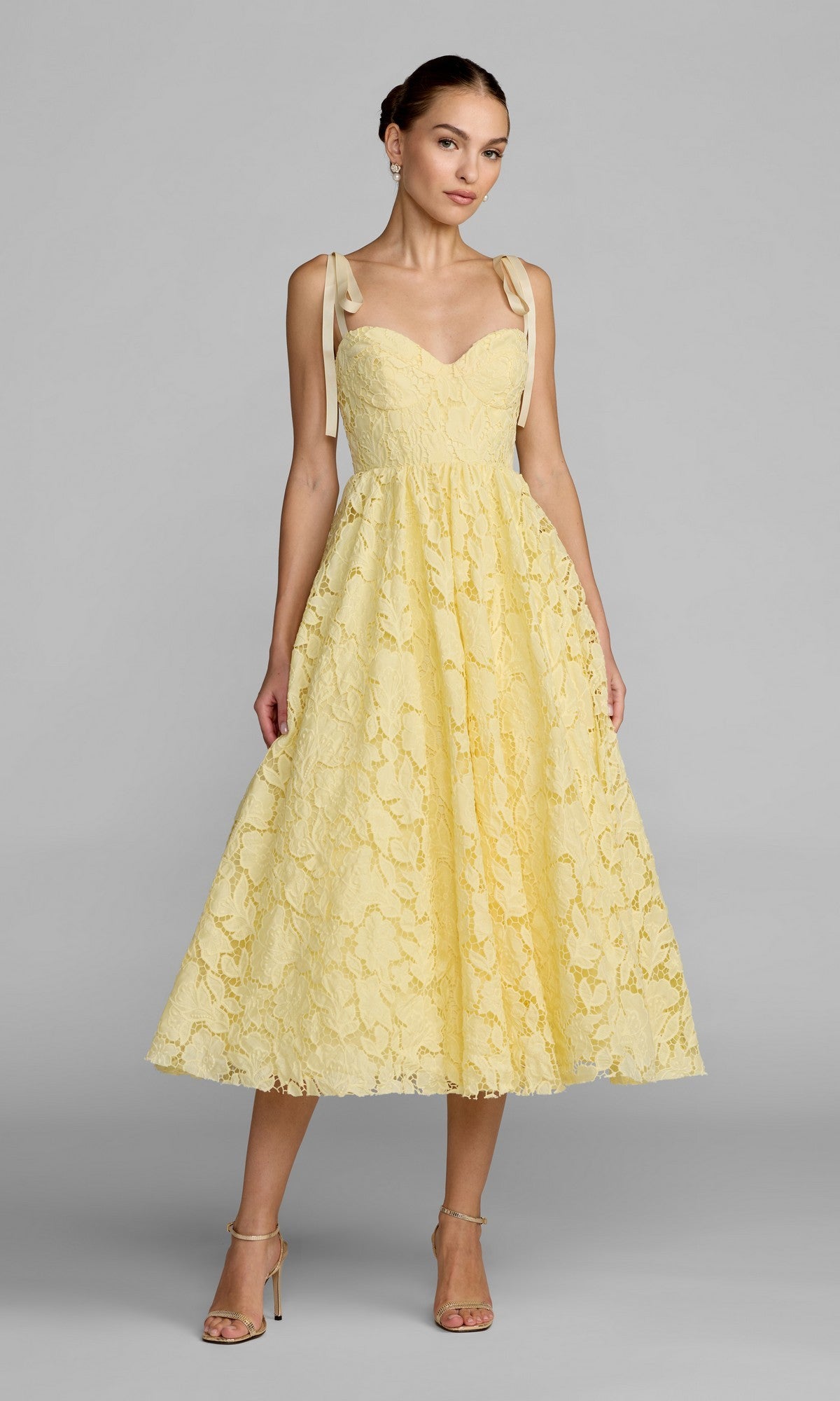 A woman wearing the Mac Duggal 56319 midi party dress in butter, featuring lace fabric and ribbon shoulder straps, stands against a plain gray background with neutral makeup and her hair styled in a neat updo.