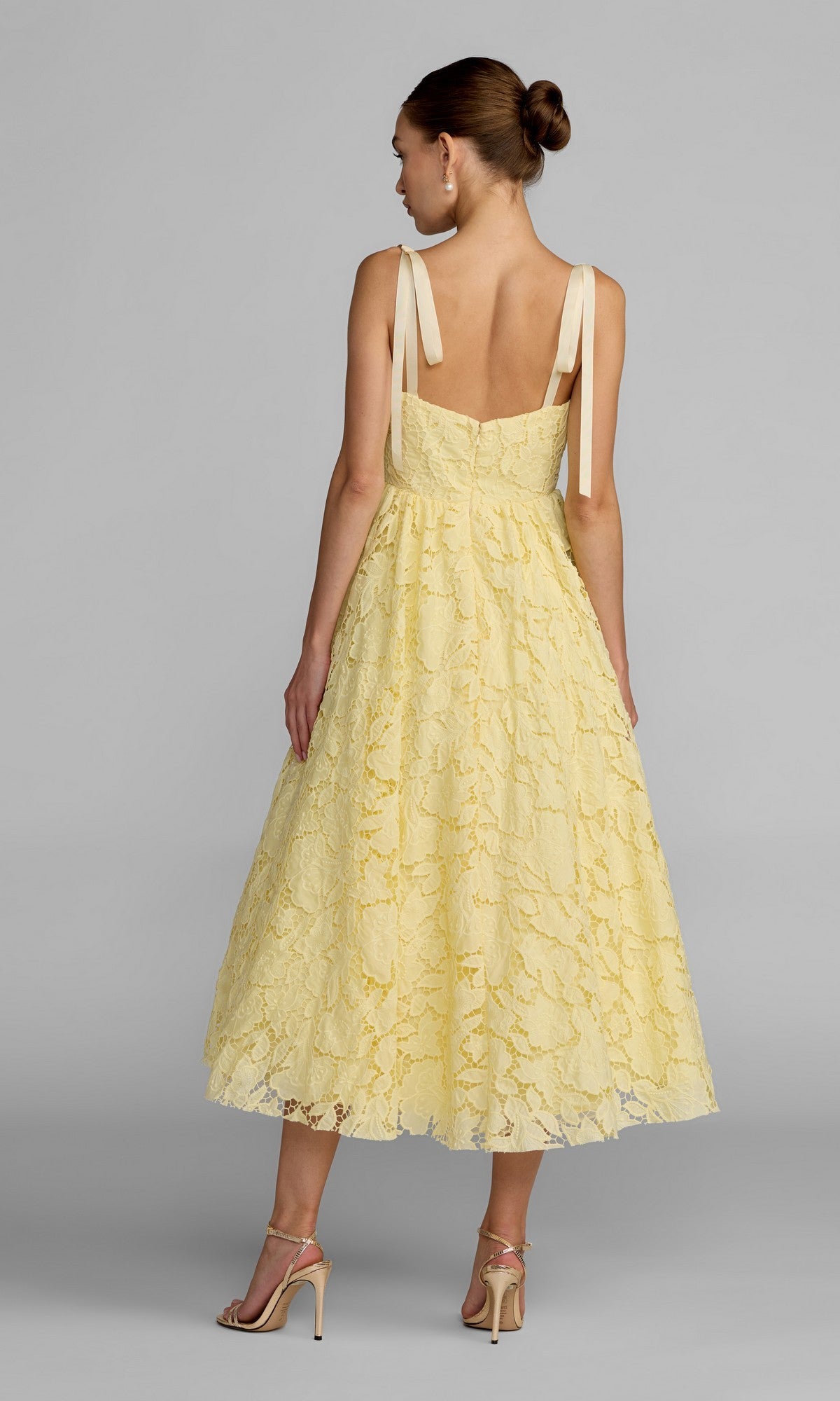 A woman stands with her back to the camera wearing the Mac Duggal 56319 Midi Party Dress—a yellow lace midi dress featuring delicate shoulder ties—paired with strappy heels, set against a plain gray background.