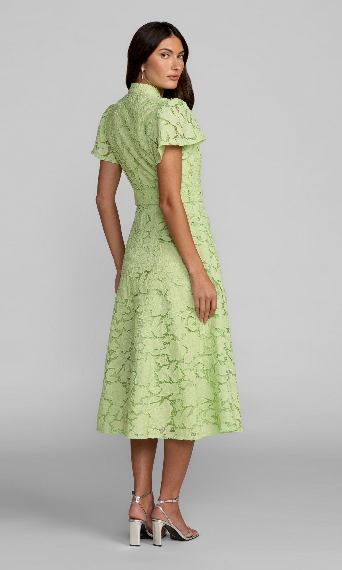 A woman is standing and facing away, wearing the Midi Party Dress: Mac Duggal 56320 in pastel green with lace embroidery and short sleeves, paired with silver high-heeled sandals against a plain gray background.