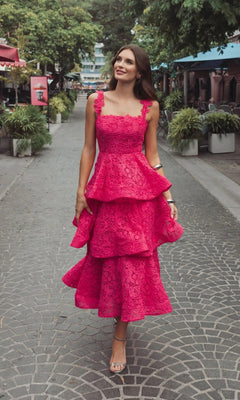 A woman walks outdoors on a paved path, surrounded by greenery and outdoor seating, wearing the sleeveless Mac Duggal 56322 Midi Party Dress with tiered lace embroidery and heels.