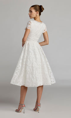 A woman stands with her back turned, looking over her shoulder against a neutral background, wearing the Midi Party Dress: Mac Duggal 56324—a white lace party dress with short sleeves and a tea-length, fit-and-flare midi silhouette. She completes the elegant look with silver high-heeled sandals.